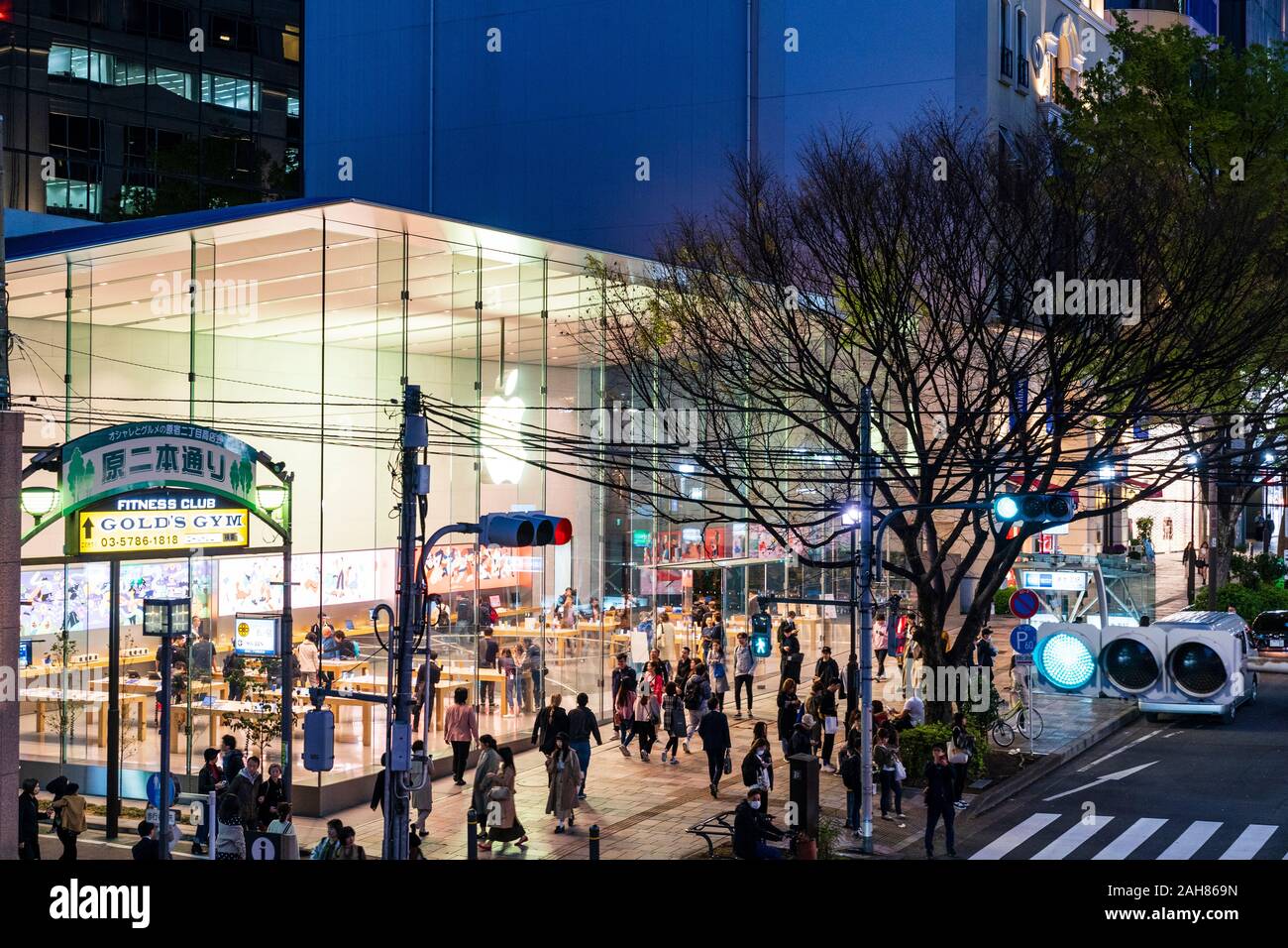 Exterior of the glass facade of the Apple store at Omotesando, Tokyo. Night time, pavement busy with shoppers, green traffic light in foreground. Stock Photo