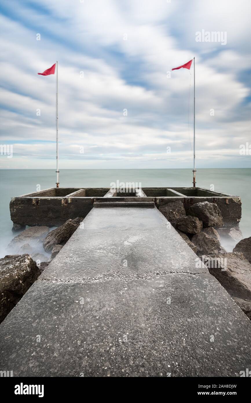 Symmetrical wide angle view of a rocky pier extending to a concrete dock flanked by two red flags Stock Photo