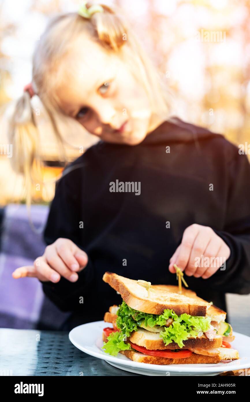 Cute adorable caucasian blond kid girl enjoy eating hamburger with ham and fresh vegeatables at cafe on bright sunny day outdoors. Fast food children Stock Photo