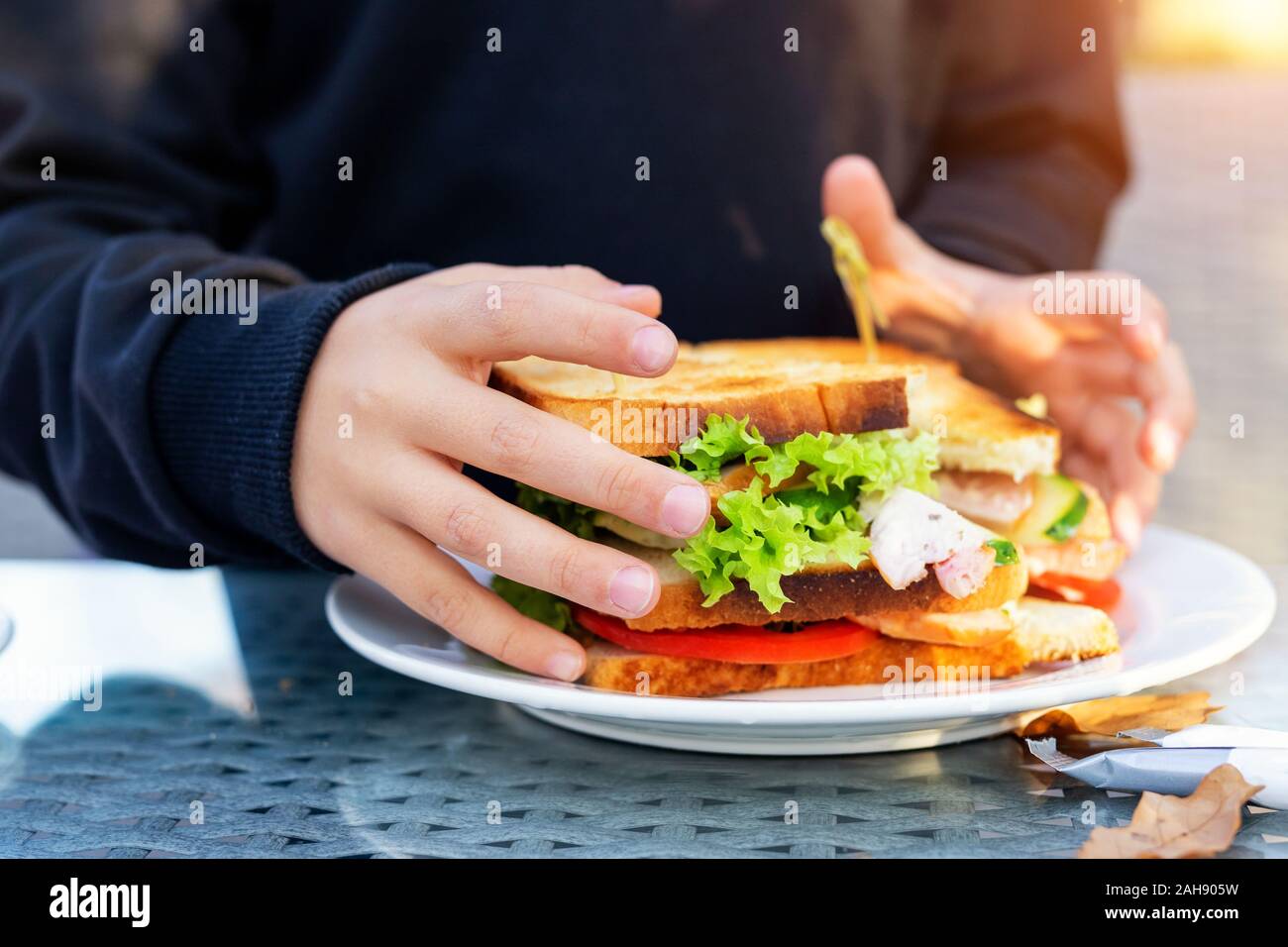 Close-up kid girl hand eating hamburger with ham and fresh vegeatables at cafe on bright sunny day outdoors. Fast food children unhealthy diet. Snack Stock Photo