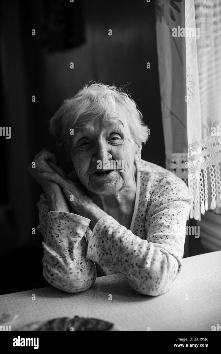 An old grandmother is sitting at a table in her house. Black and white photo. Stock Photo