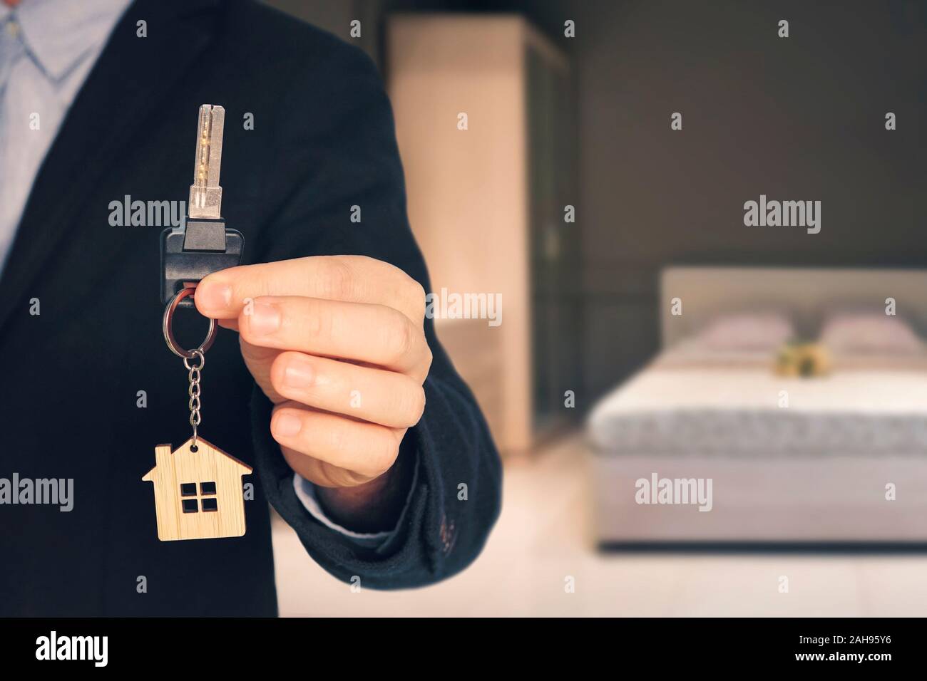 Man shows keys with keychain in form of a little house into a new flat. blurred background of the bedroom room with a modern interior. Stock Photo