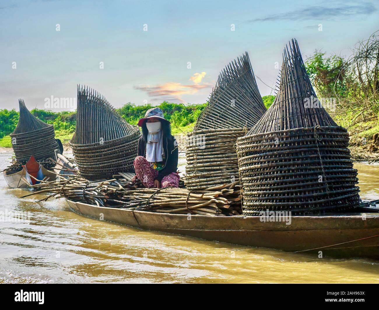 A Cambodian woman sits in a wooden boat being towed down a river. The boat is filled with homemade fishing trap. Stock Photo