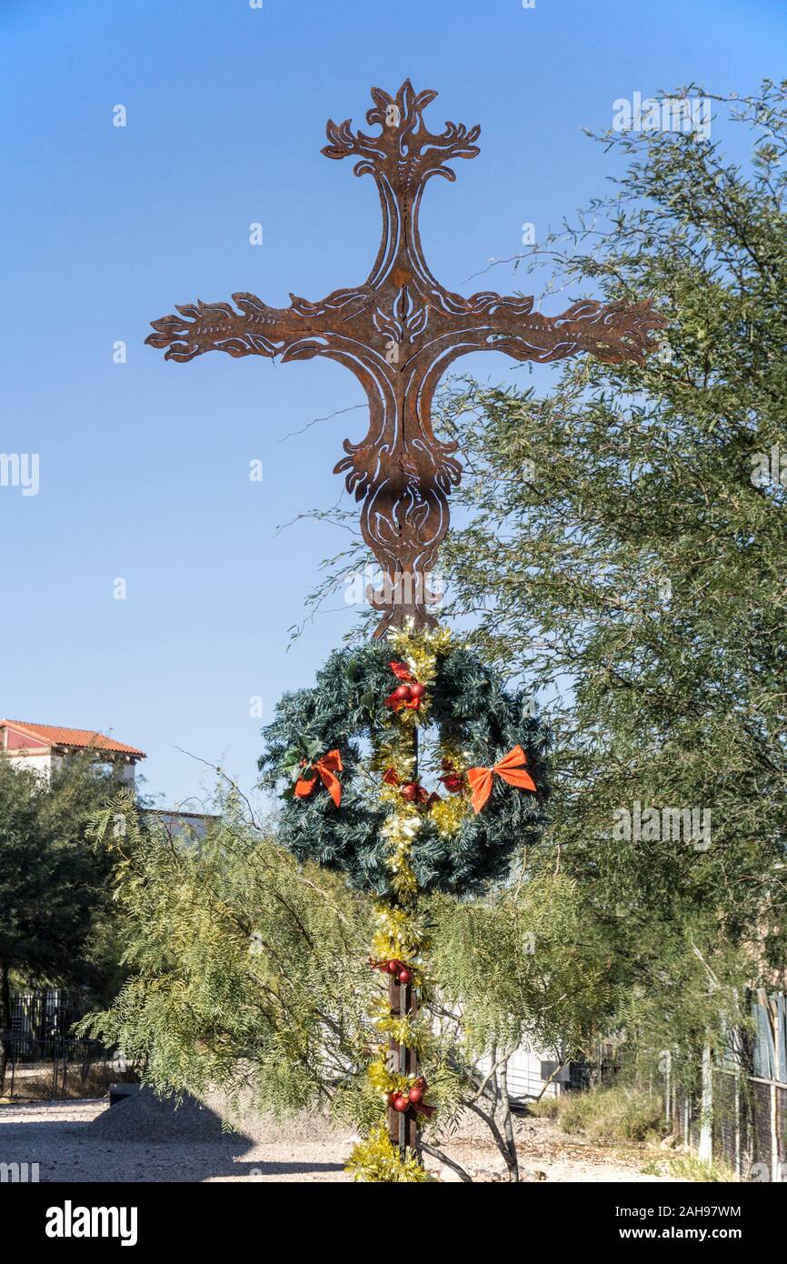 view ornamental rusting iron cross mounted on pole in garden San Cosme Roman Catholic Church garlanded with wreath for the Christmas season in Tucson Stock Photo