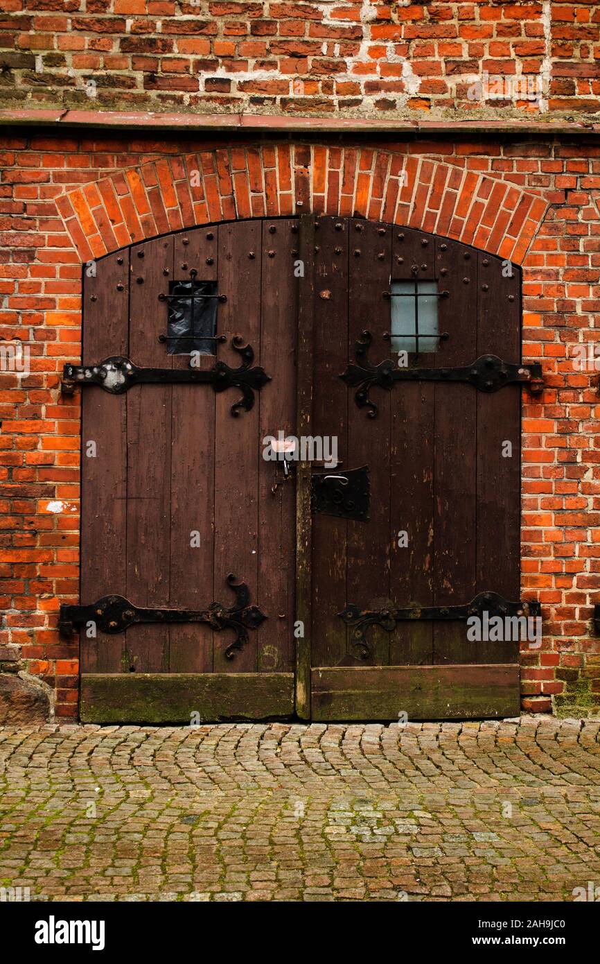 Two winged wooden doors with iron-wrought supports, of an old brick building with cobblestone ground at the foreground. Stock Photo