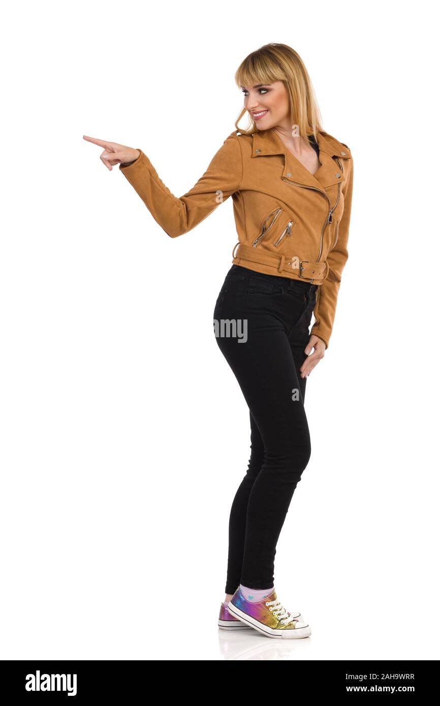 Smiling young woman in brown suede leather jacket, black jeans and sneakers is standing, looking away over the shoulder and pushing something or point Stock Photo