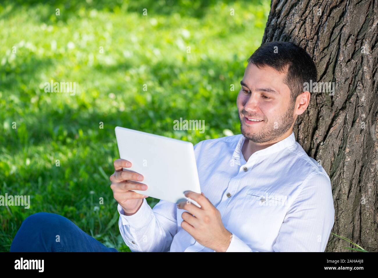 Man using tablet computer under tree in park on sunny day. Handsome guy looking at tablet screen. Happy smiling man sitting on green grass leaning aga Stock Photo