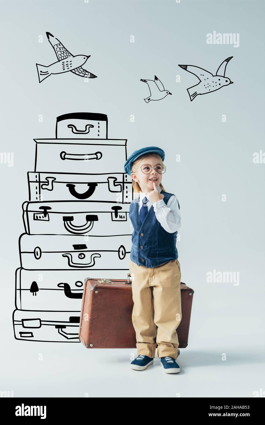 dreamy kid in retro vest and cap holding suitcase and looking away on grey background with travel bags and flying birds fantasy illustration Stock Photo
