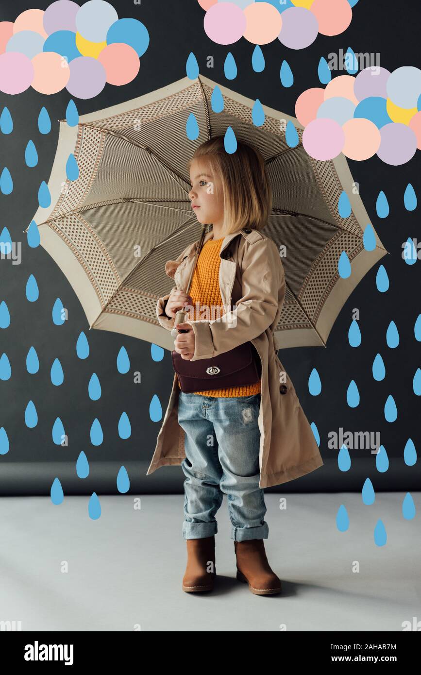 serious child in trench coat and jeans holding umbrella under fairy rain and looking away Stock Photo