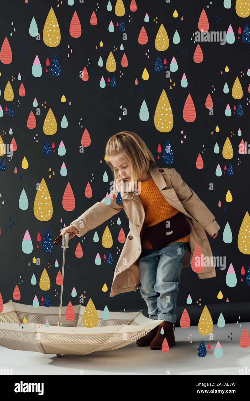 serious child in trench coat and jeans holding umbrella under fairy colorful rain Stock Photo