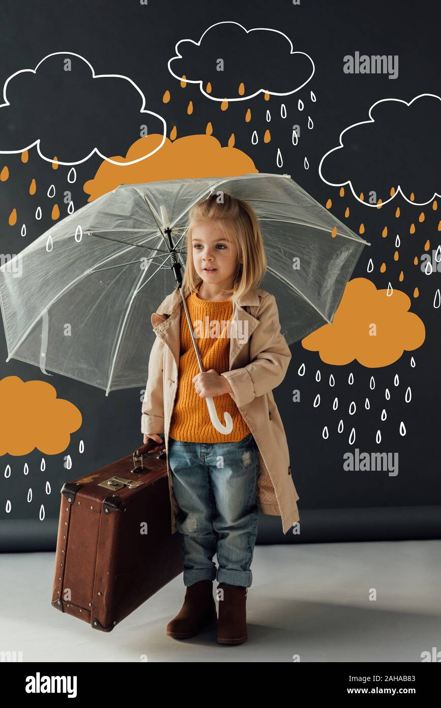 child in trench coat and jeans holding umbrella and leather suitcase under fantasy rain on black background Stock Photo