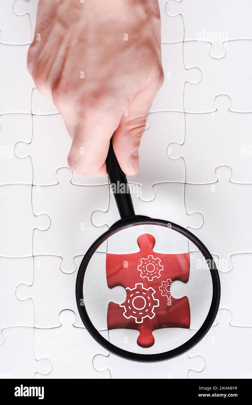 top view of man holding magnifying glass near jigsaw with gears