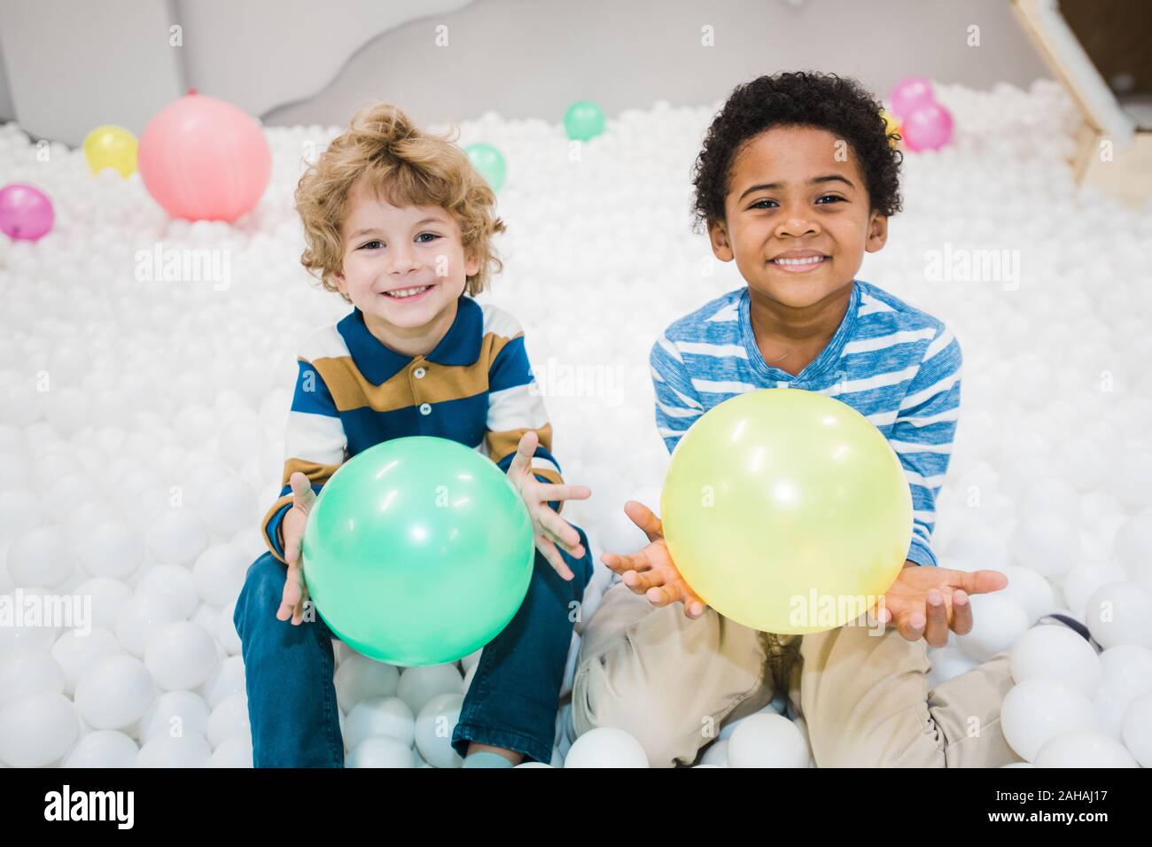 Cute cheerful intercultural little boys in striped shirts playing with balloons Stock Photo