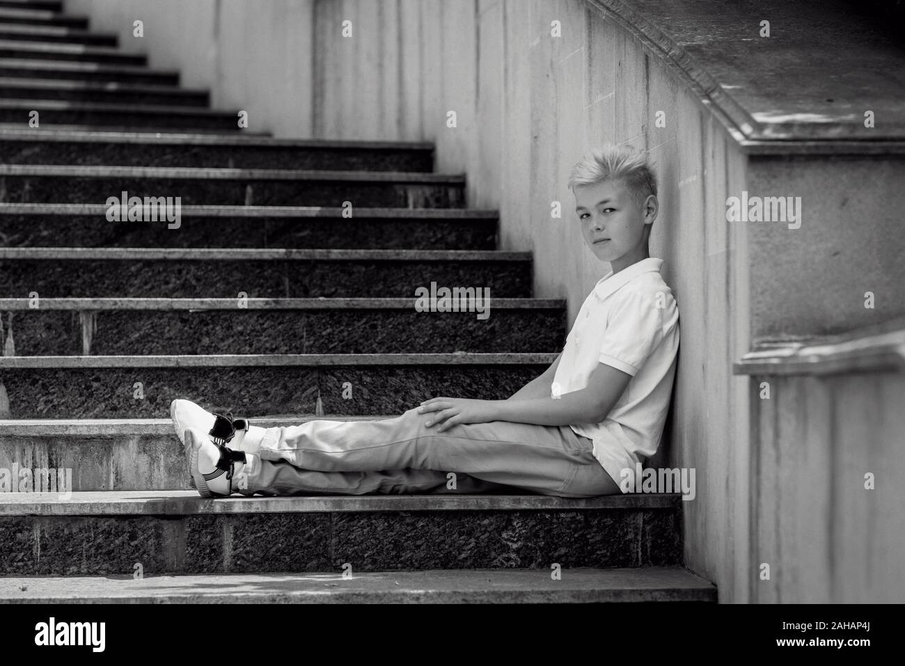 monochrome (black and white) portrait of cute handsome blonde eleven old boy (teenager) sitting on the stairs outdoor.Growing up, chilhood, separating Stock Photo