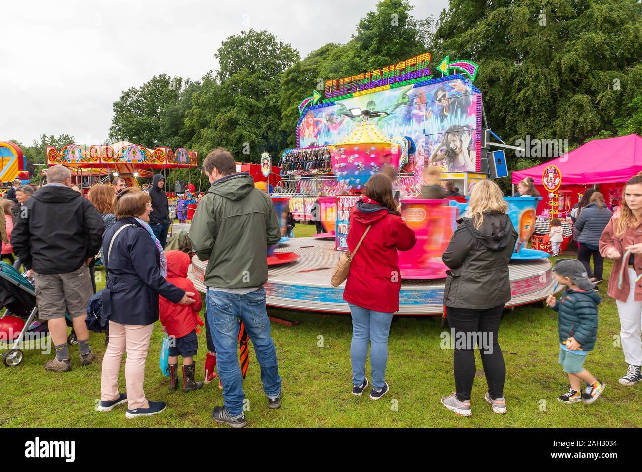 People watching the Cliffhanger fairground ride and a children's tea cups ride in wet weather in summer after the Thelwall Rose Queen procession 2019 Stock Photo