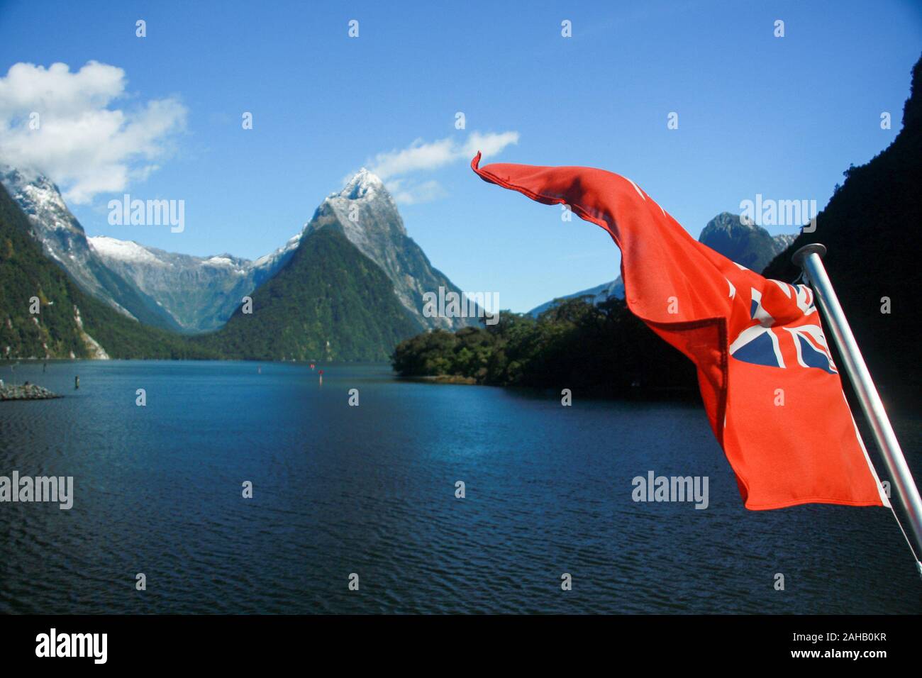 The Red Ensign, a red maritime version of the NZ flag, flutters on a sightseeing ferry boat on the calm, deep blue waters of Milford Sound in the remote Fiordland region in New Zealand's South Stock Photo