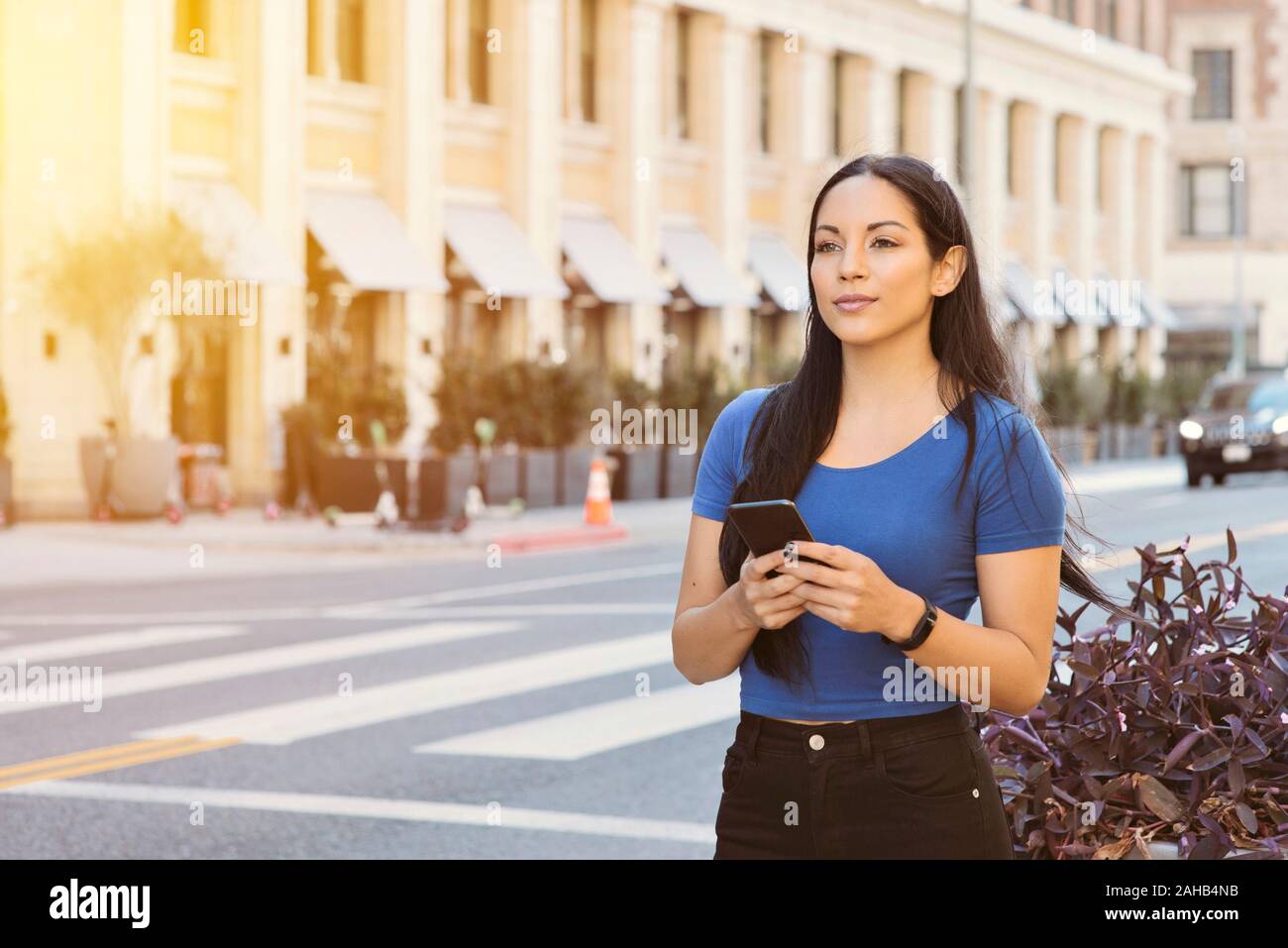 Young attractive girl awaits a ride share car in the city - Holding smart phone waiting on the sidewalk - Daytime with warm lighting Stock Photo