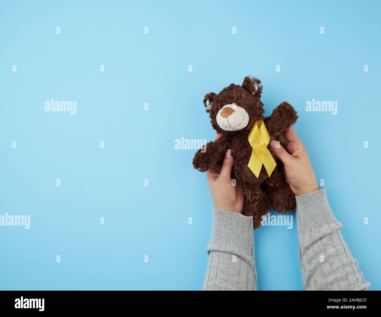 female hands hold a small brown teddy bear which holds in its paw a yellow ribbon folded in a loop on a blue background. concept of the fight against Stock Photo
