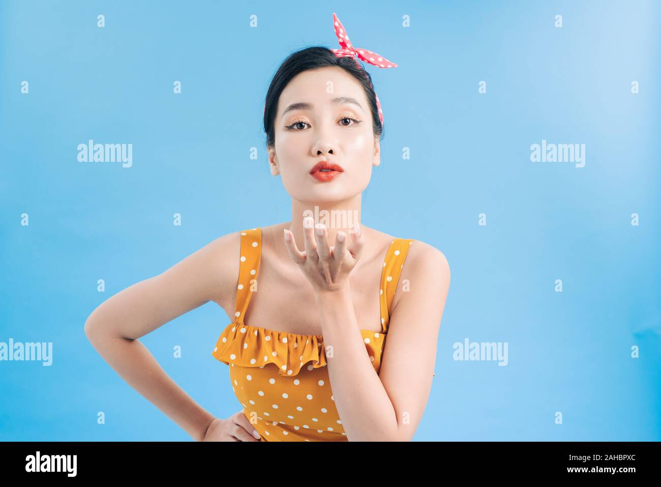 Attractive stylish young female wearing retro yellow dotted dress holding open palms and pouting lips, blowing air kisses Stock Photo