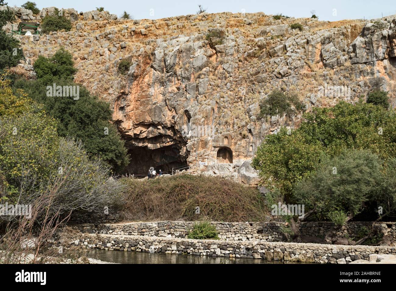 tourists exploring the cave of pan at the base of a tall cliff in the hermon stream park in israel with trees and water pools in the foreground Stock Photo