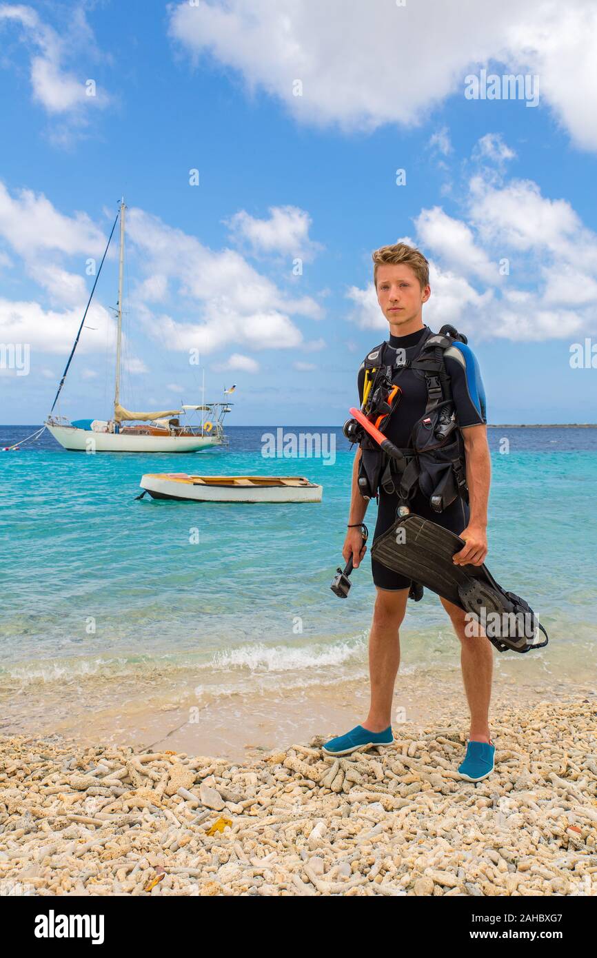 European male diver standing on beach of Bonaire with sea and boats Stock Photo