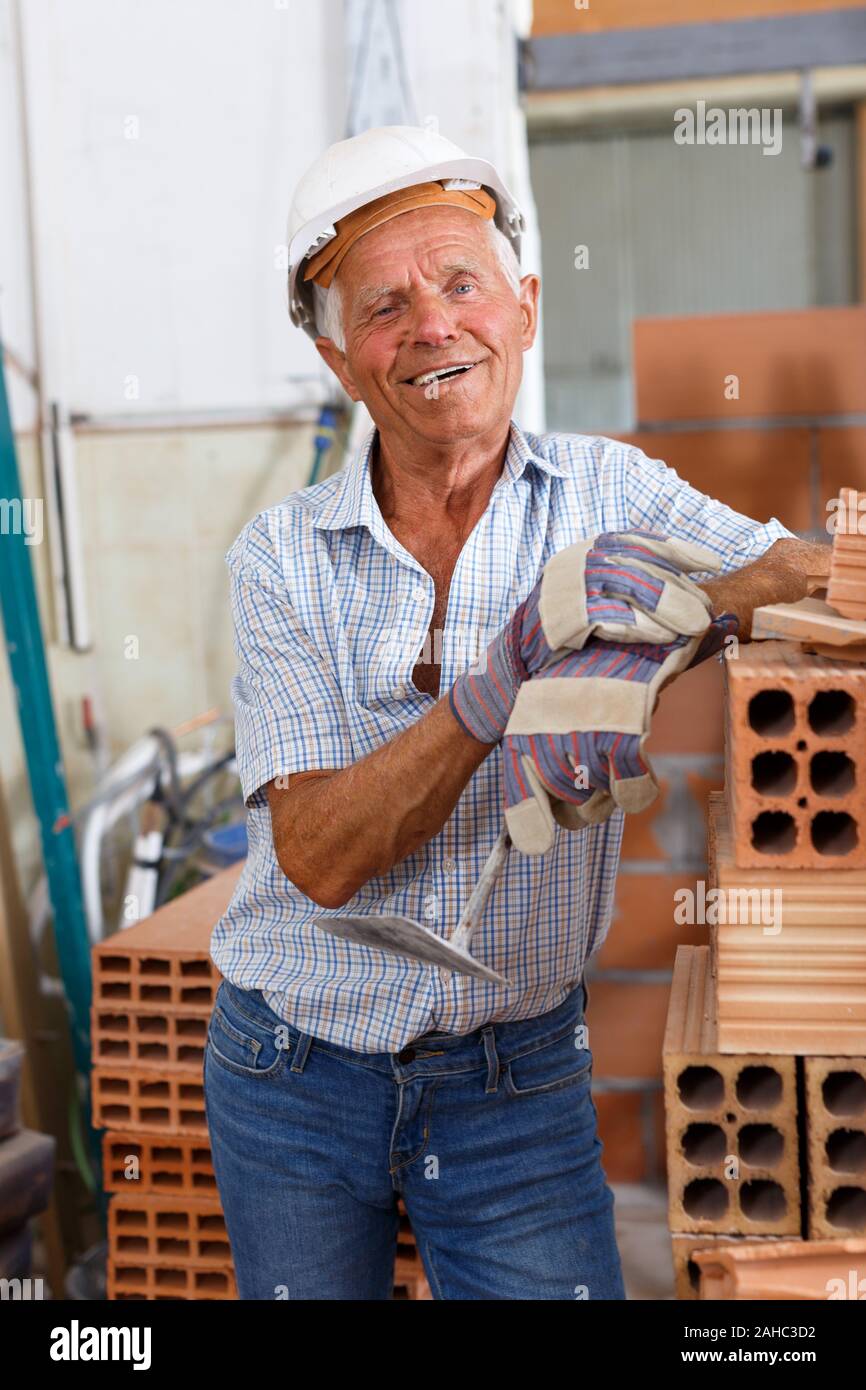 Happy experienced elderly bricklayer posing near red brick stack in building under construction Stock Photo