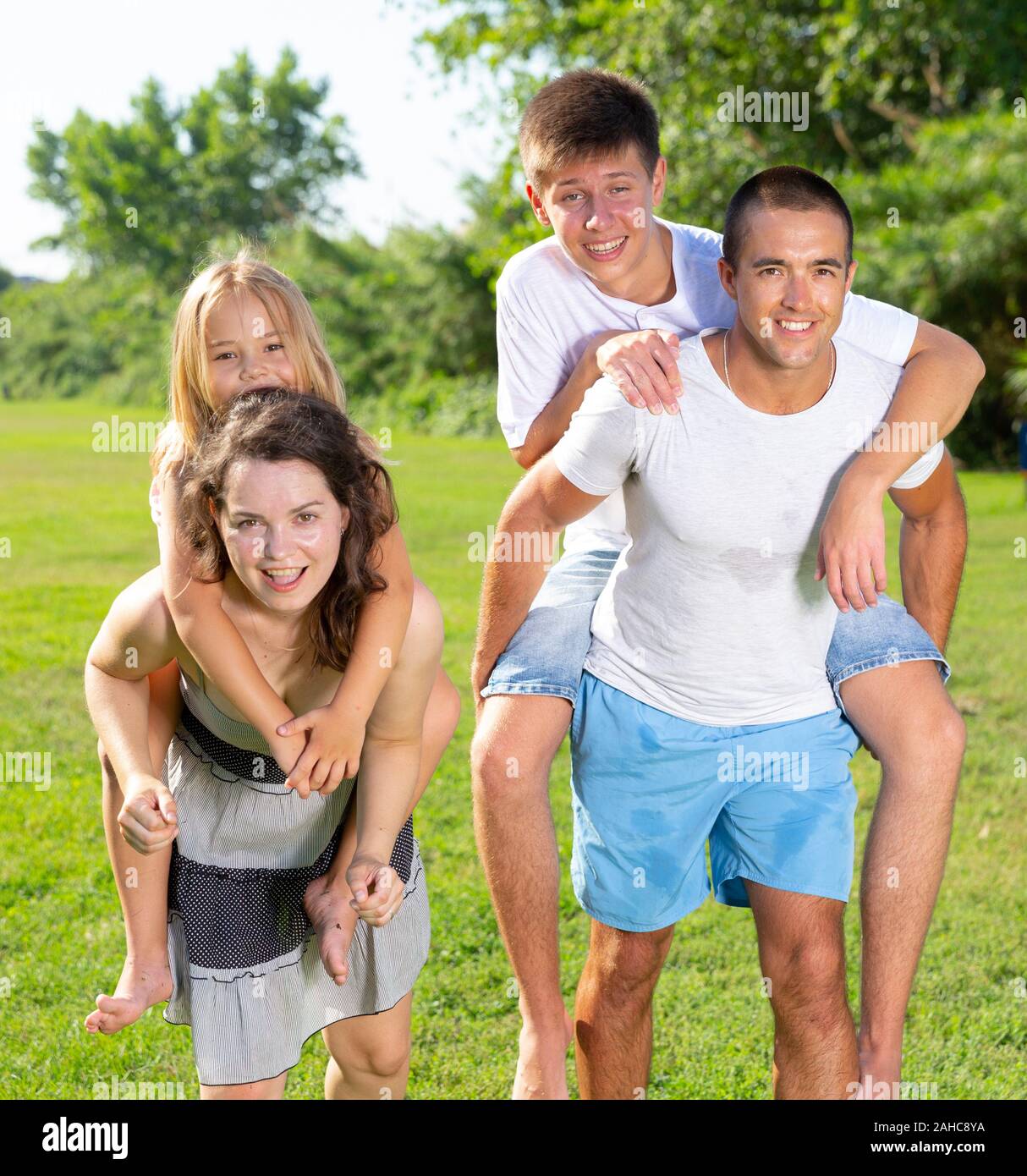 Cheerful friendly family having fun together in summer city park, parents piggybacking children Stock Photo