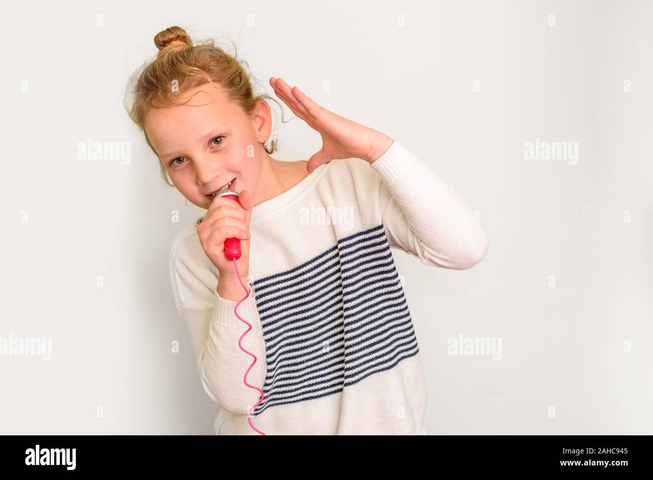 Photo of little meloman girl listens favorite playlist enjoys good sound, holds microphone and singing, wears casual clothing, on white background. Monochrome. Stock Photo