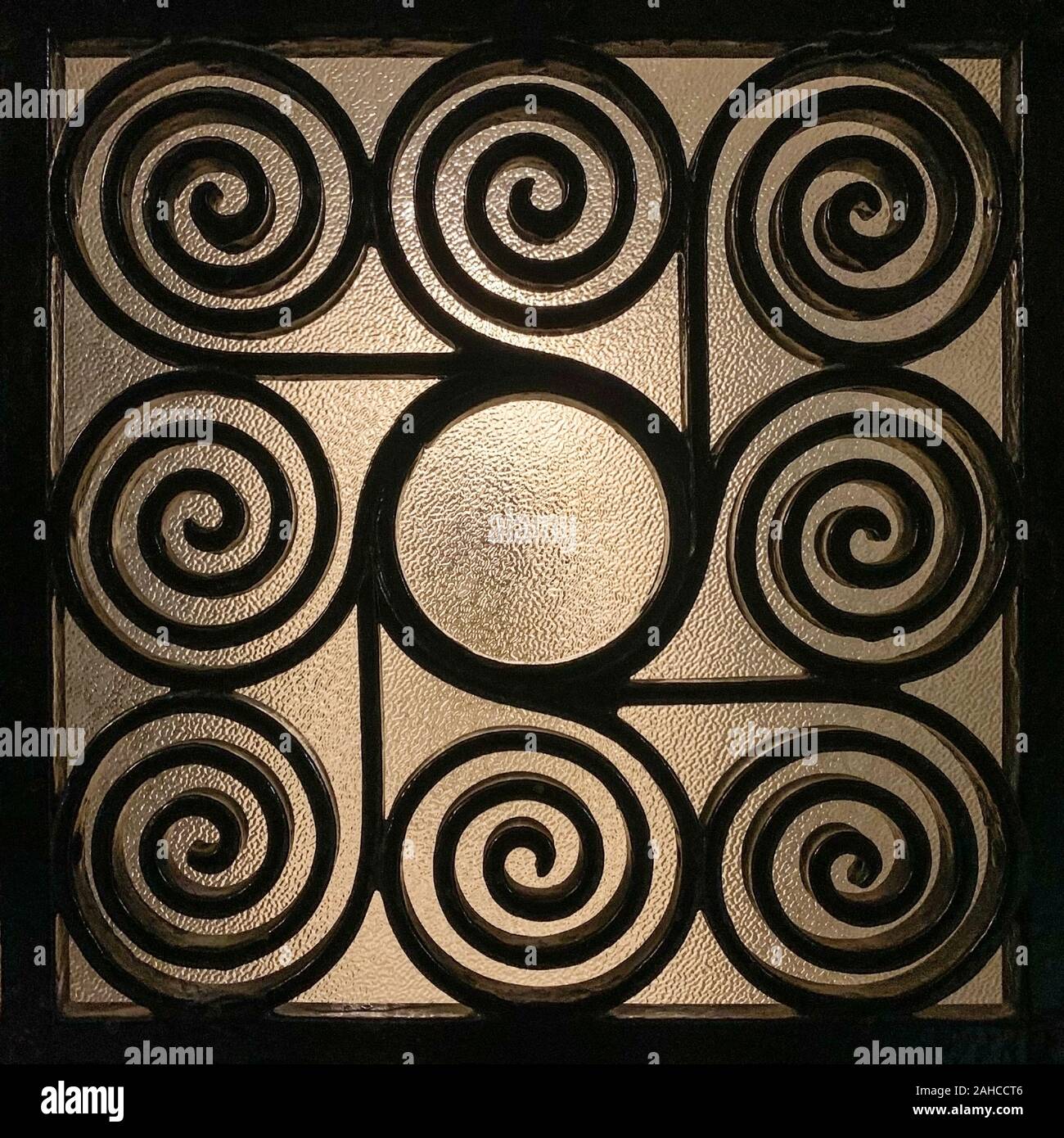 Wrought Greek iron grille, ornamented with elaborately twisted spirals and with frosted glass in the background, that is slightly illuminated. Stock Photo