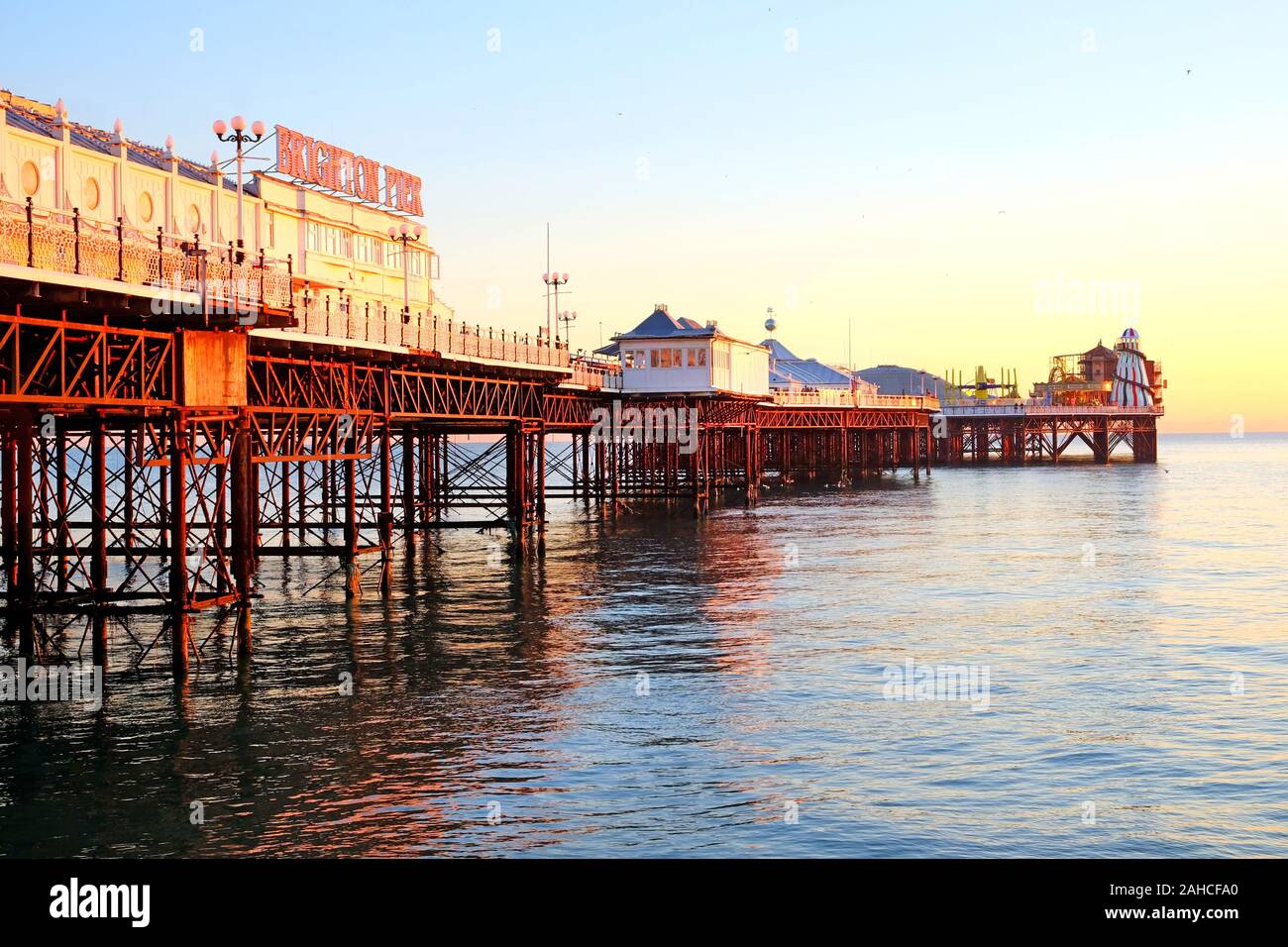 the full length of Brighton pier, the pier has an orange glow from the sun setting, the mane of the pier is on the left and the fairground is at the e Stock Photo