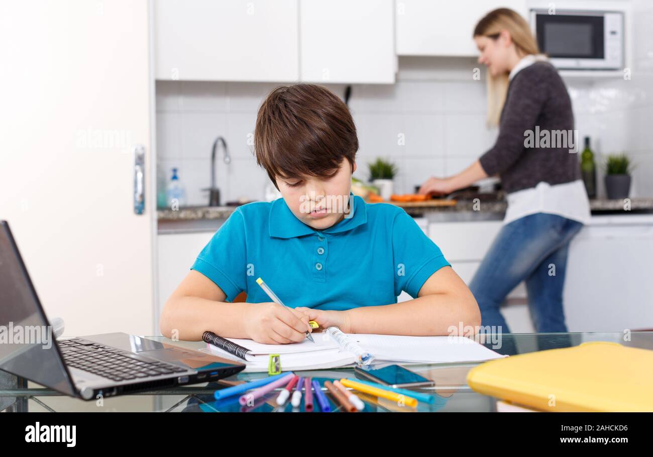 School age boy doing homework at kitchen while his mother preparing ...