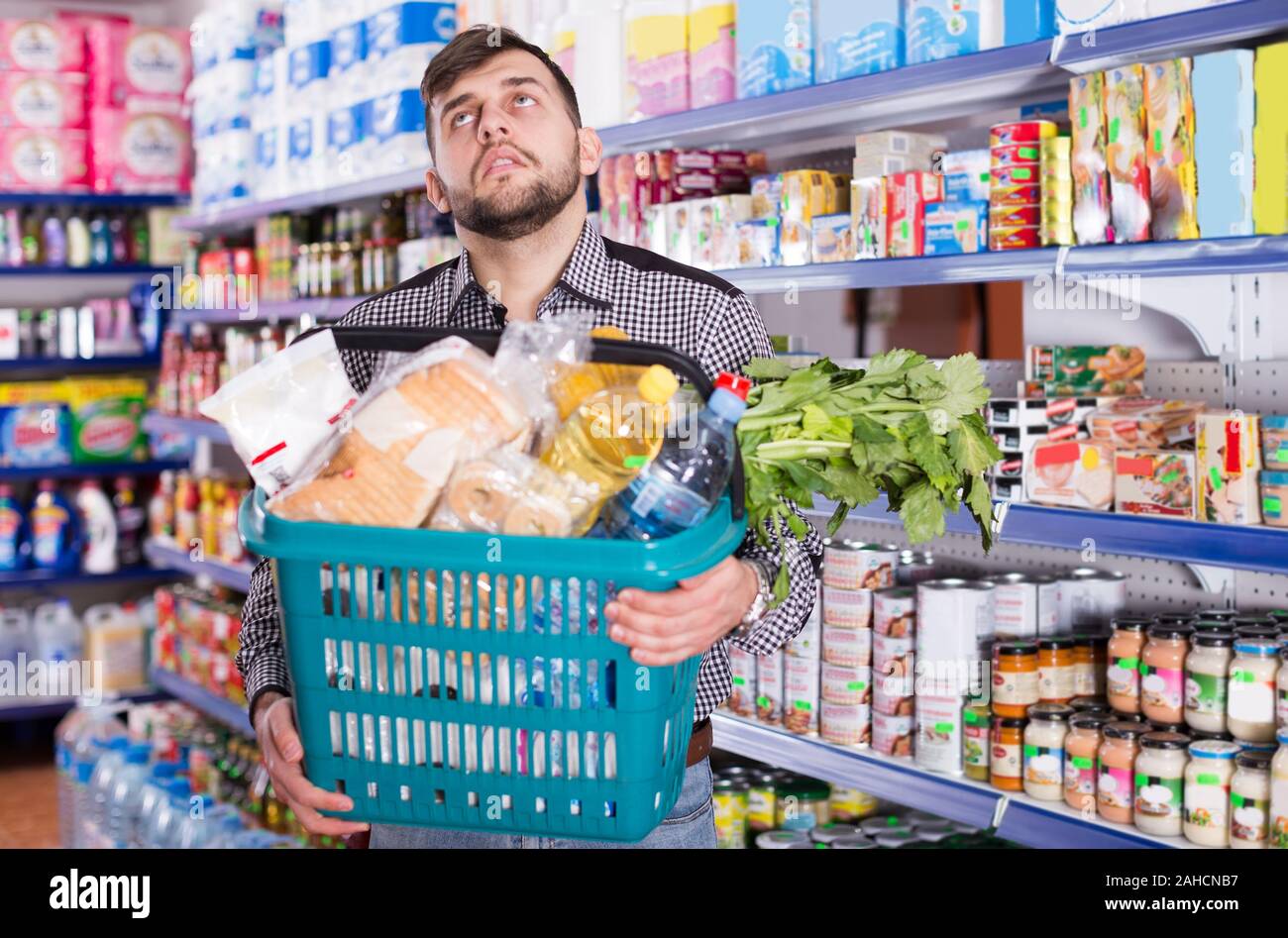 Tired young male with heavy shopping basket filled food products in supermarket Stock Photo