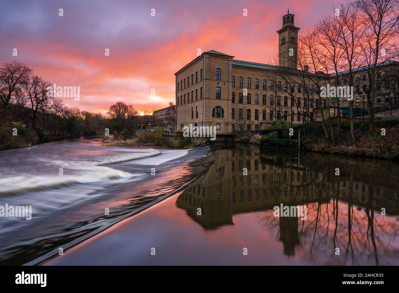 A vibrant sunrise frames New Mill at the Saltaire UNESCO World Heritage Site, with the distinctive architecture reflected in the River Aire. Stock Photo