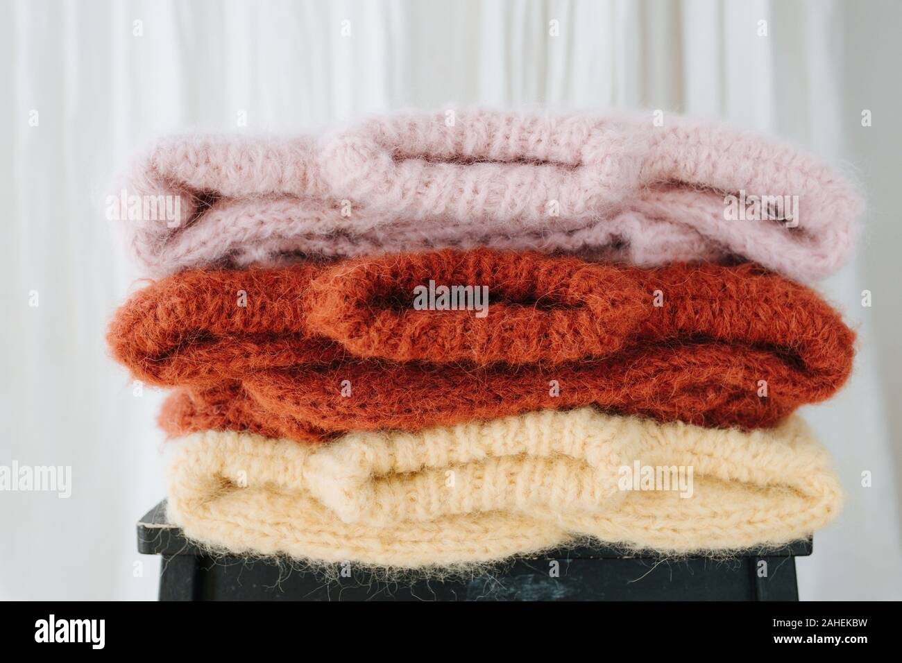 Three folded multi-colored sweaters lie on a stool in front of a curtain. Stock Photo