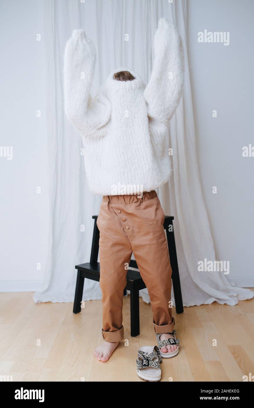 Funny little girl struggling to put on white fluffy knitted sweater. Stock Photo