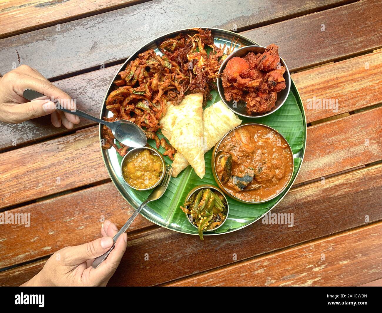 Man using fork and spoon to eat banana leaf dish, consists of fried chicken, curry seafood, lady fingerspotatoes and canai Stock Photo