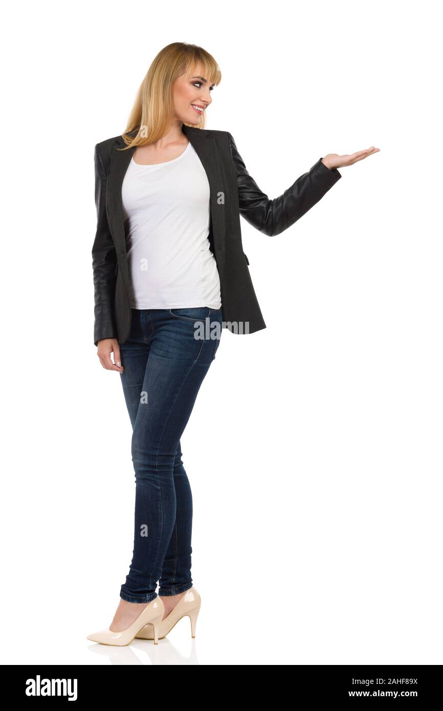 Smiling young woman in black jacket, blue jeans and high heels is standing, looking away over the shoulder, holding hand raised and presenting. Full l Stock Photo