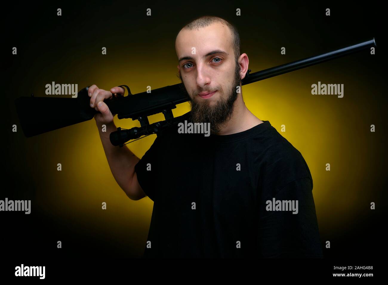 Portrait of a man holding a shotgun riffle over his shoulder. Handsome guy shot against dark yellow background Stock Photo