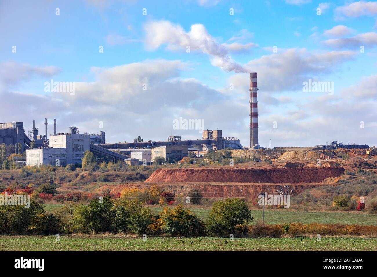 The pipe of the cement plant rises above the quarry of clay and limestone against the background of a slightly cloudy blue summer sky. Stock Photo