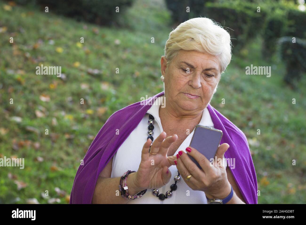 Senior woman dialing on screen touch cellphone in the park. Elder blonde lady with purple sweater over shoulders texting on smart phone outdoors Stock Photo