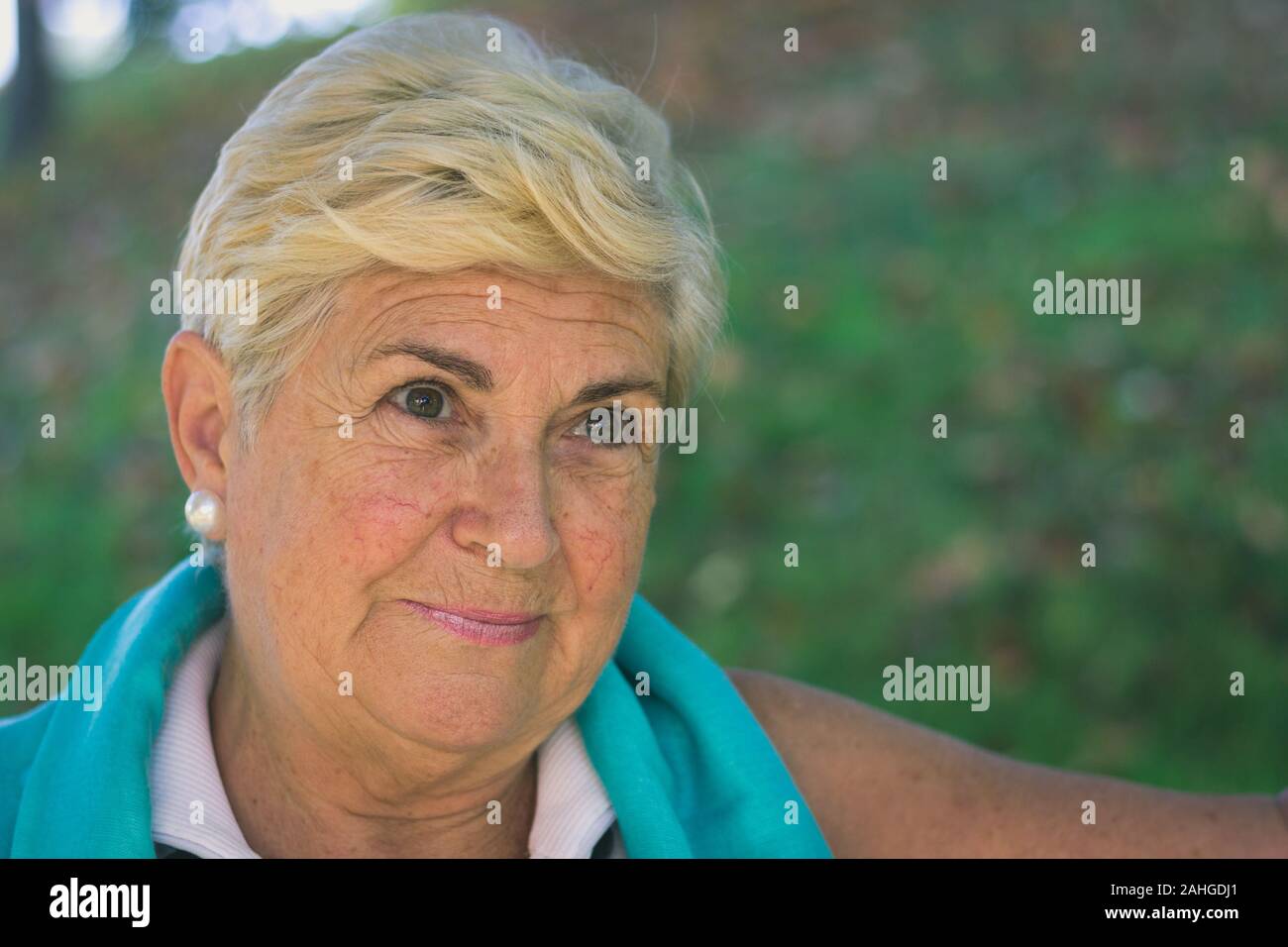 Pensive elder lady portrait in park. Senior blonde woman smiling with optimistic look. Closeup of mature person with sleeveless white shirt Stock Photo