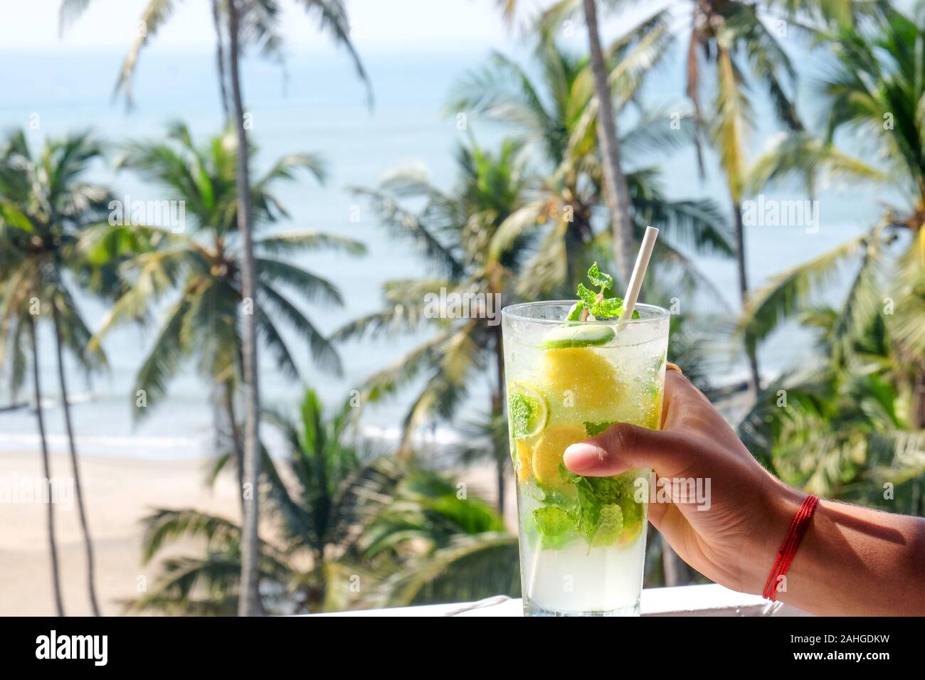 A young Asian man’s hand holding a virgin mojito cocktail brightly shining in the sunshine, behind is a tropical scene of palm trees, a sandy beach an Stock Photo