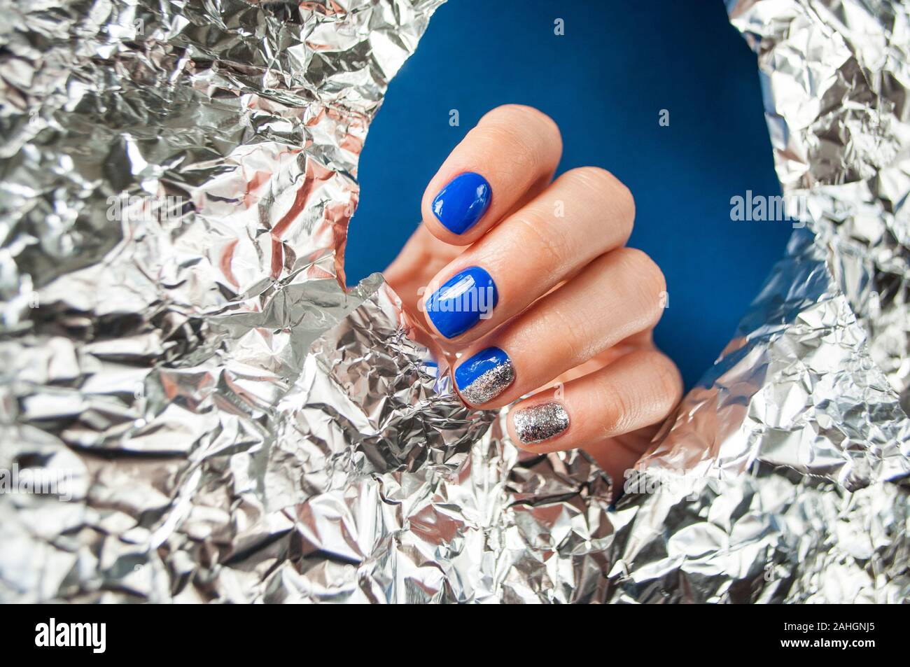 Young woman's hand with beautiful manicure blue on blue background with metallic shiny paper. 2020 color trend. Stock Photo