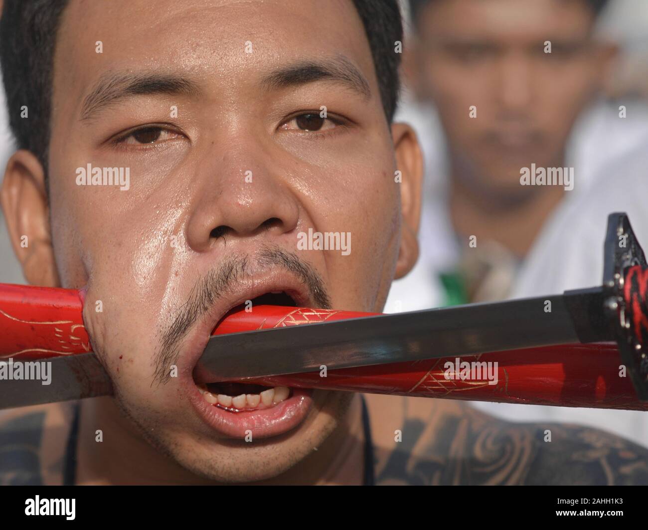 Young Thai Chinese Taoist devotee pierces his cheek with the blade and scabbard of an Asian sword during the Nine Emperor Gods Festival. Stock Photo