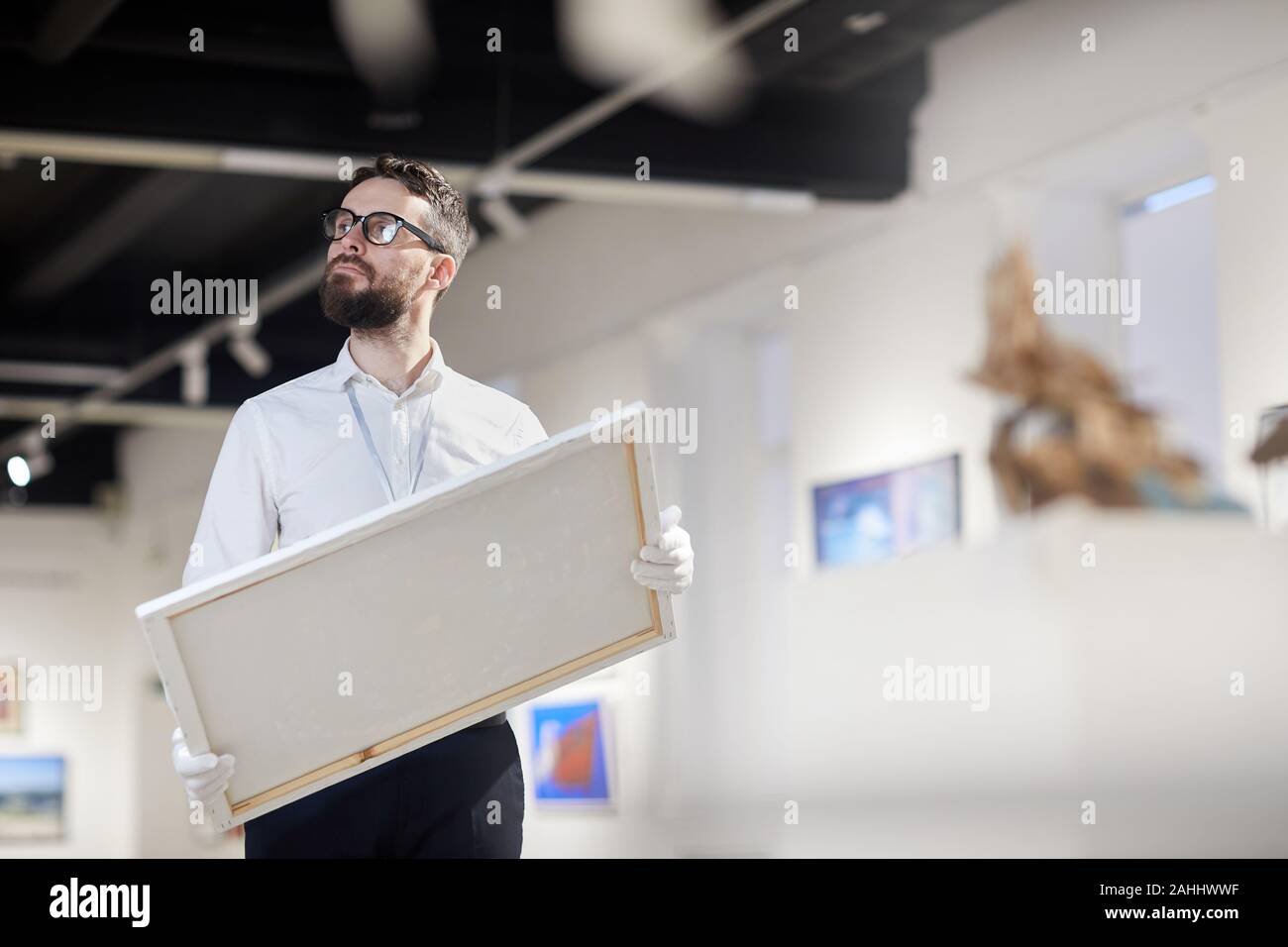 Waist up portrait of bearded art gallery worker holding painting while
