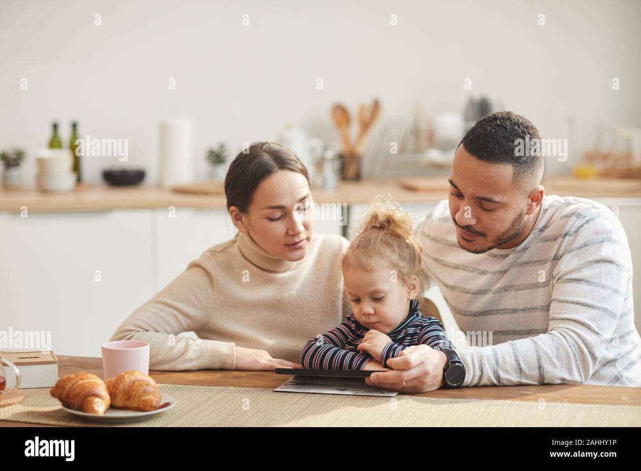 Warm-toned portrait of modern family with cute little daughter using digital tablet together while sitting at wooden table in cozy kitchen, copy space Stock Photo