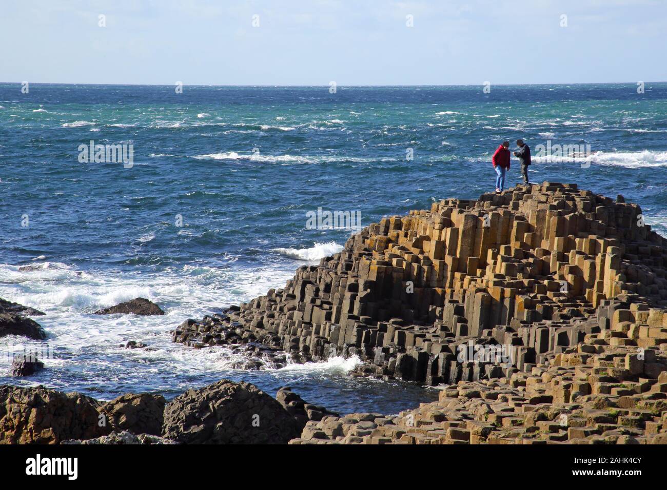 Massive basalt columns of the Giant's Causeway with two men standing on the summit taking photographs with their mobile phones, blue sky and sea, Coun Stock Photo