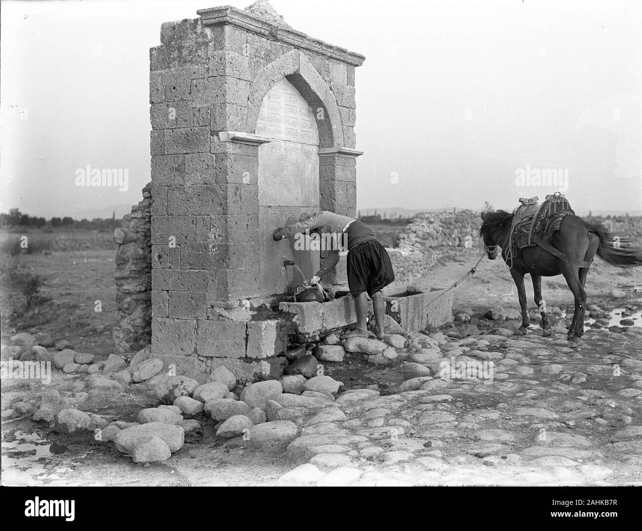 Turkey Ottoman countryman in traditional dress at a public 'Menzil' fountain with his horse, filling a water jug. These Menzils were located outside settlements and served caravans, travellers, farmers and their beasts. The fountains had inscriptions in verses, with the last line giving information about the construction date, either directly or in Ebced calculation. Copy from a dry glass plate, originating from the Herry W. Schaefer collection. Stock Photo