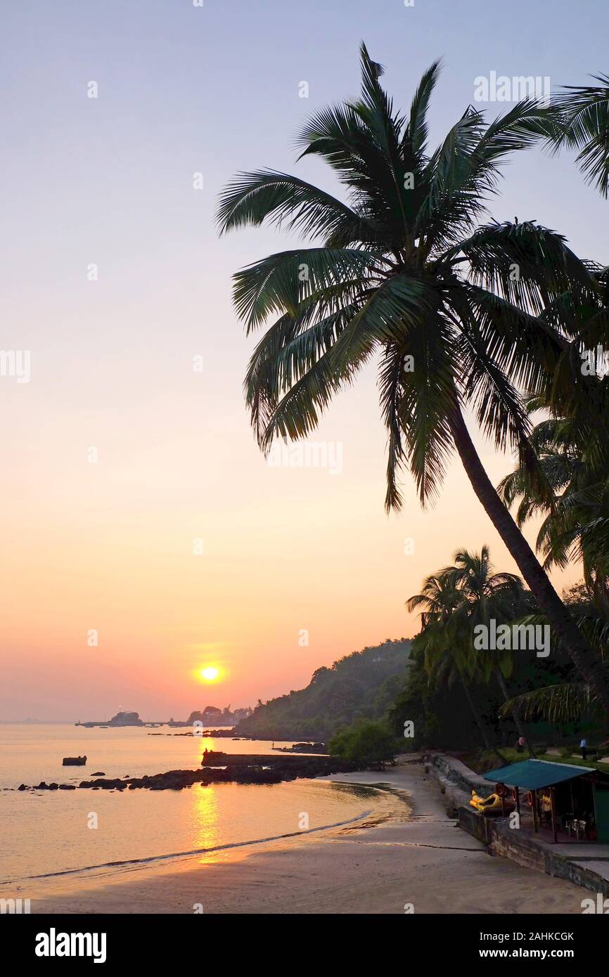 A tropical bay with a calm sea and palm trees, Red glow from the setting sun over Viaguinim Beach, Panaji, Goa, India, vertical format Stock Photo