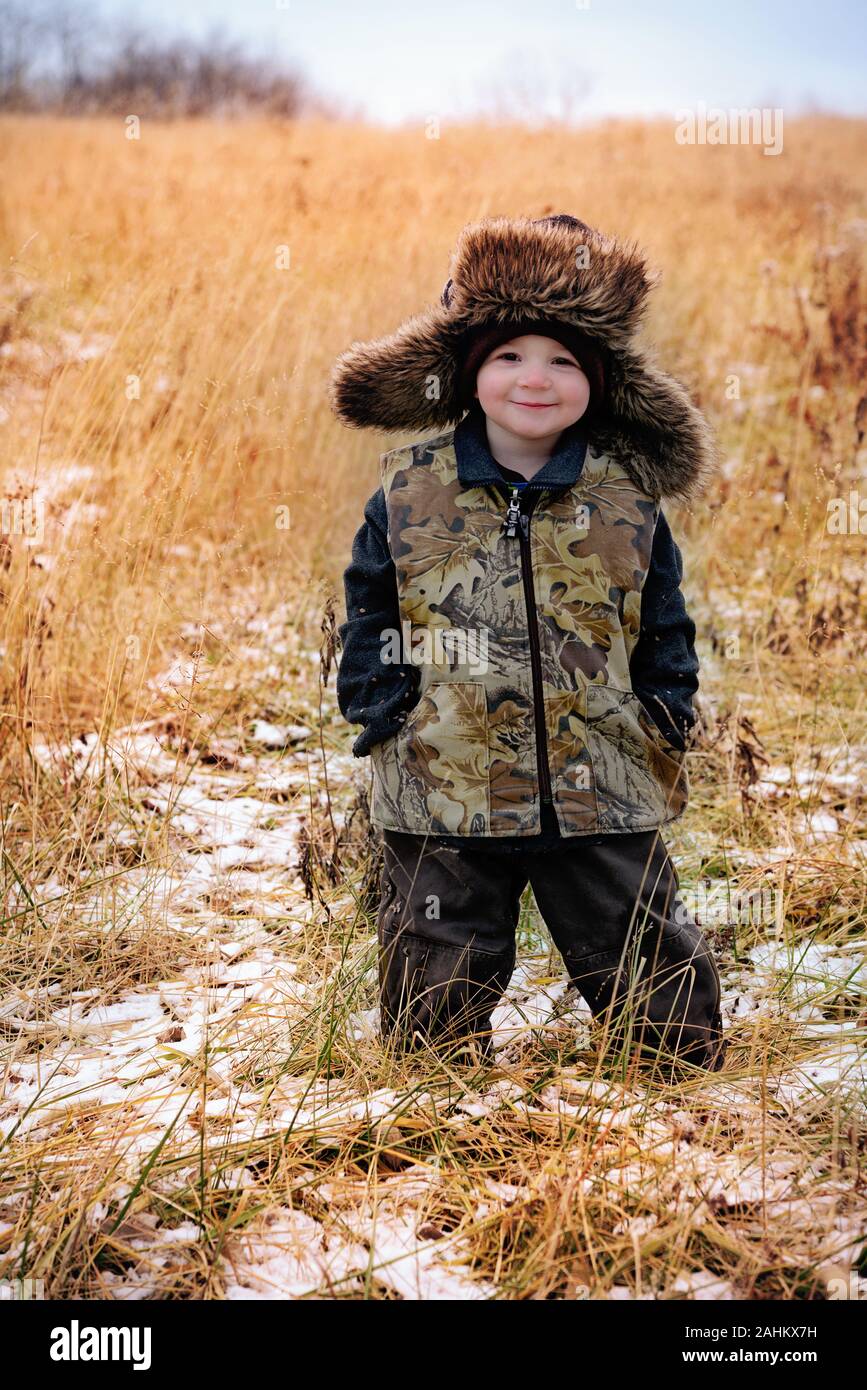 little boy wearing camoflauge vest and  fur-lined trapper hat in a country field Stock Photo